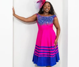 The quintessential ensemble for race day elegance. Woman wearing a hot pink midi dress with blue floral bodice embroidery and striped hem, paired with a matching fascinator.